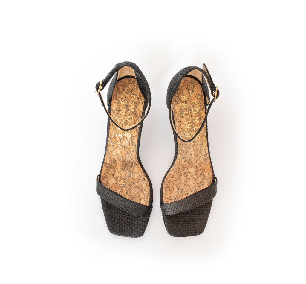 Low Stiletto in Black Raffia, ankle strap with metal buckle. Mid-heel in recycled wood pulp, covered with black raffia.