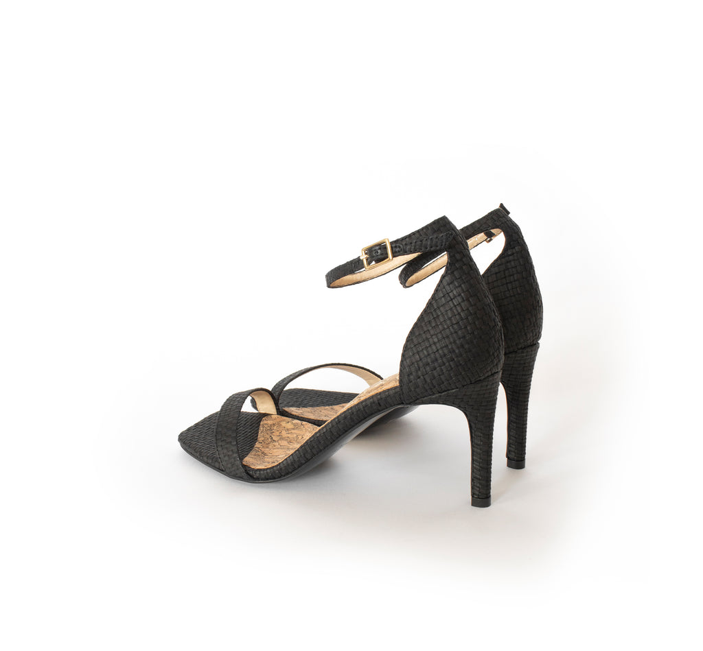 Low Stiletto in Black Raffia, ankle strap with metal buckle. Mid-heel in recycled wood pulp, covered with black raffia.