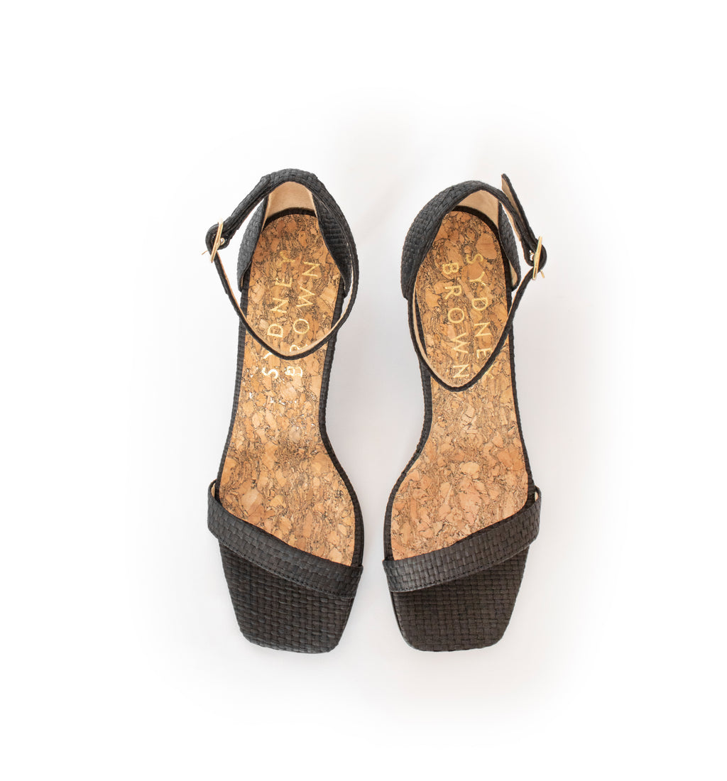 Low Stiletto in Black Raffia, ankle strap with metal buckle. Mid-heel in recycled wood pulp, covered with black raffia.