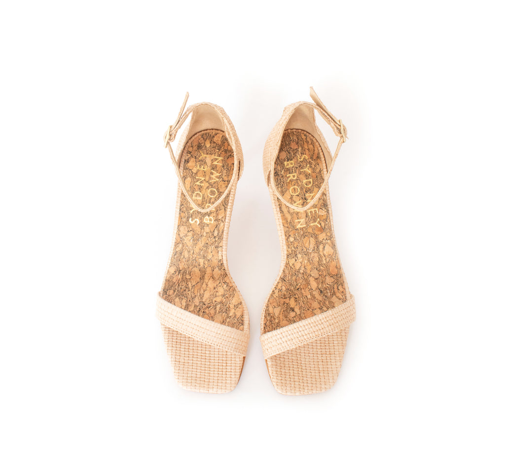 Low Stiletto in natural raffia, ankle strap with metal buckle. Mid-heel in recycled wood pulp, covered with natural raffia.